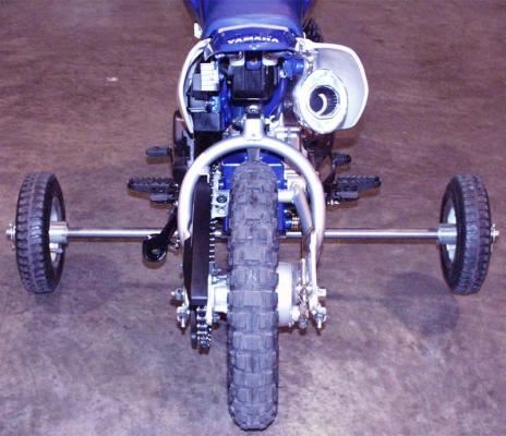 The training wheels attach to the underside of the motorcycle frame beneath the engine.